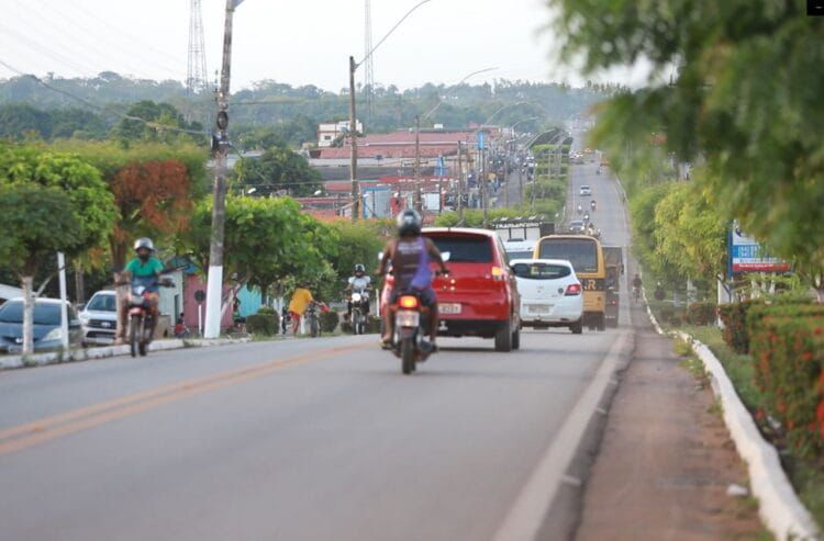 Rua movimentada com carros e motos subindo uma ladeira arborizada em uma cidade brasileira.