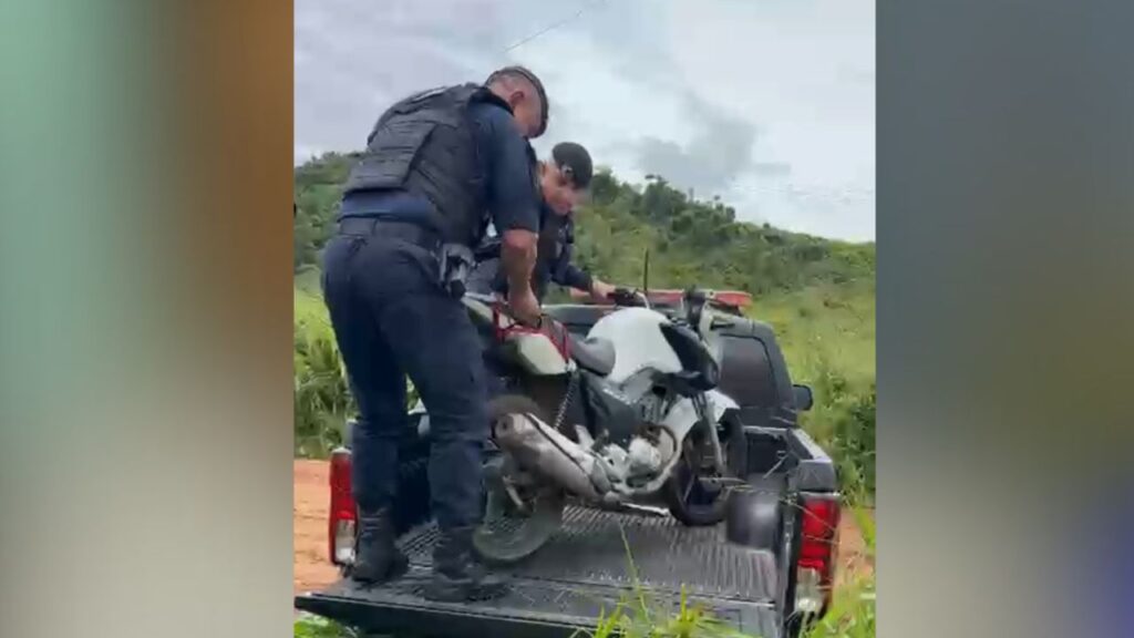 Dois policiais ou agentes de segurança carregando uma motocicleta branca na caçamba de uma caminhonete em área rural.