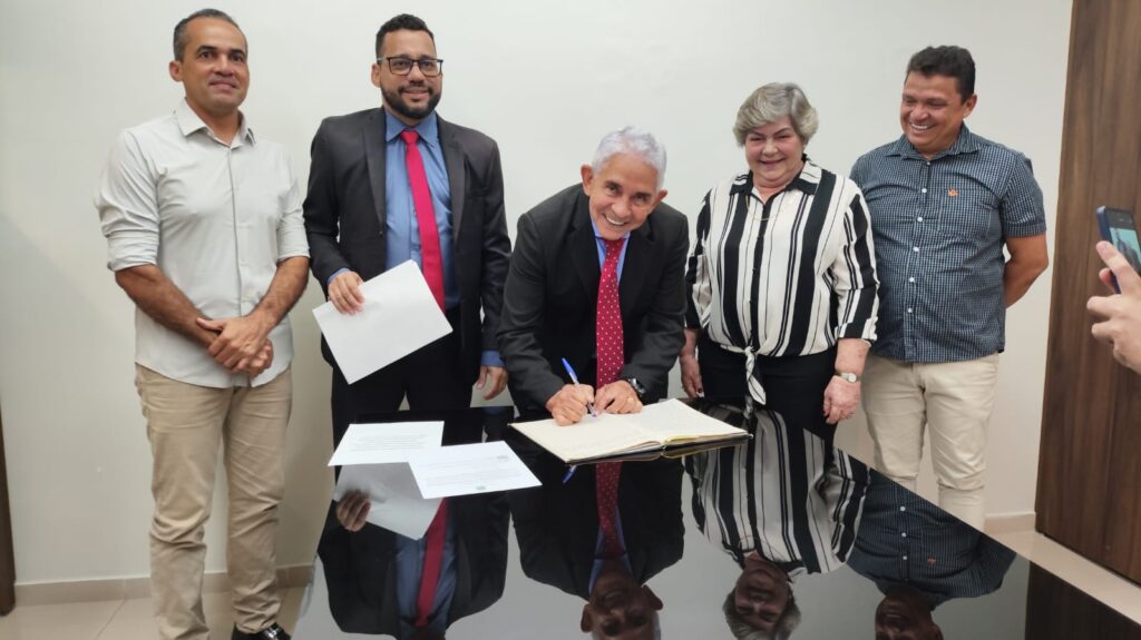Homem sorridente de terno assinando um documento em uma mesa preta, com outras pessoas ao redor.