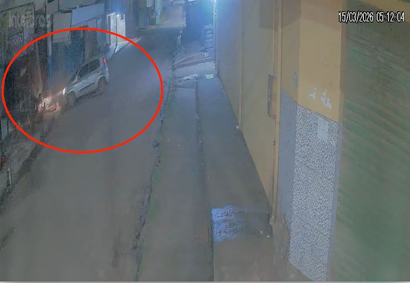 Imagem de câmera de segurança mostrando um carro claro com faróis acesos em uma rua escura ao amanhecer, realçado por um círculo vermelho.