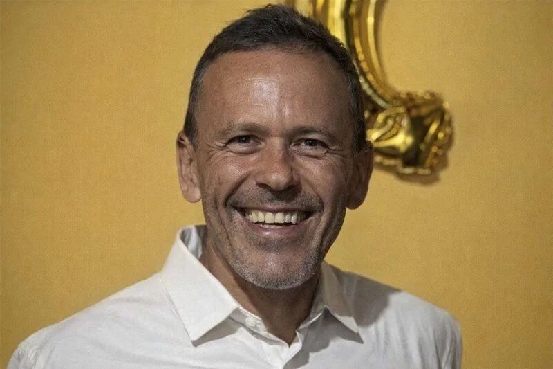 Homem de meia-idade sorrindo, usando camisa branca, contra um fundo amarelo.