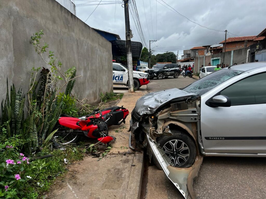 Acidente de trânsito grave entre carro prata e moto vermelha em rua residencial, com viaturas policiais ao fundo.