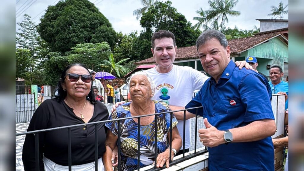 Duas mulheres e dois homens sorrindo para a câmera em ambiente externo com árvores e uma multidão ao fundo.