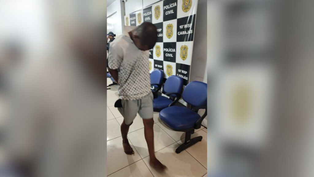 Homem com as mãos para trás e cabeça baixa em delegacia, com banner da Polícia Civil ao fundo.