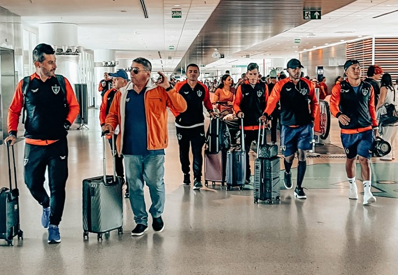 Um grupo de homens, incluindo atletas e membros da equipe, caminhando com malas em um aeroporto.