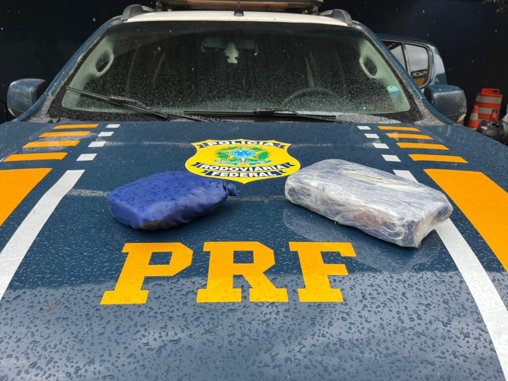 Dois pacotes de material ilícito sobre o capô molhado de uma viatura da Polícia Rodoviária Federal (PRF), com o emblema e letras da corporação.