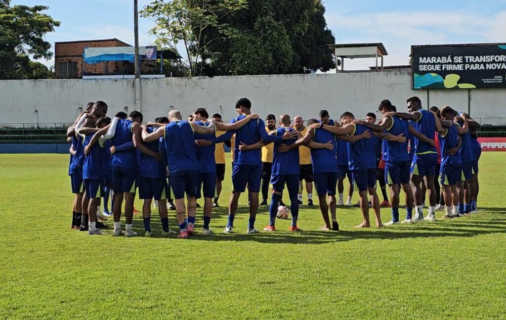 Um grupo de jogadores de futebol em círculo, abraçados, em campo gramado.