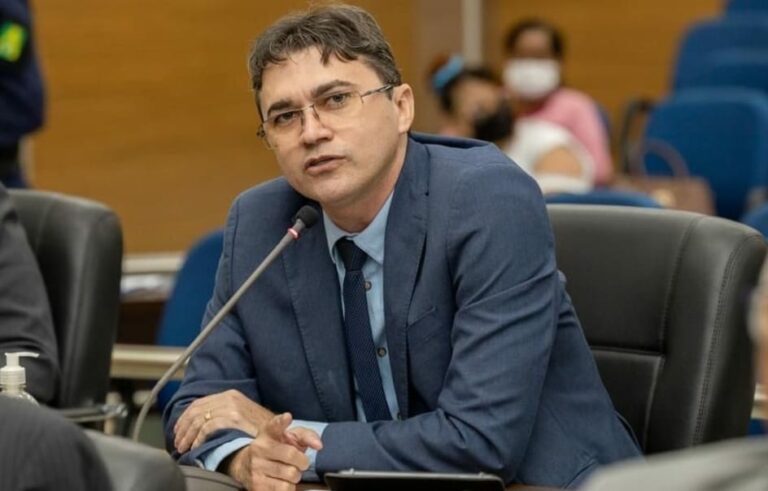 Homem de terno azul e óculos falando em microfone durante uma reunião formal.