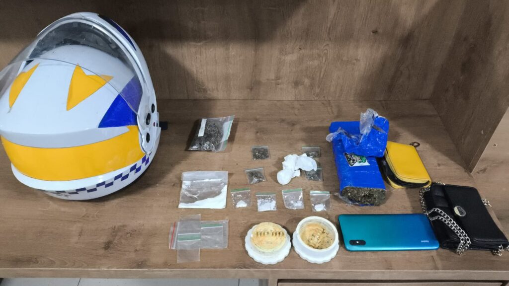 Visão aérea de diversas drogas ilícitas, incluindo maconha e pó branco, em embalagens plásticas, com um capacete, um celular e moedores em uma mesa de madeira.