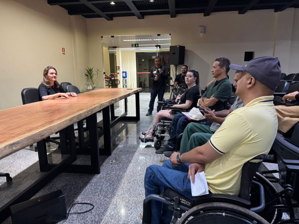Grupo de pessoas em cadeiras de rodas participando de uma reunião com uma mulher sentada em uma mesa de madeira.