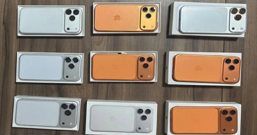 Nove cases ou modelos de smartphones com câmera tripla, em cores prata e laranja, dentro de caixas brancas sobre uma mesa de madeira.