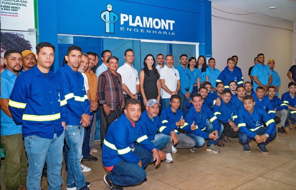 Grupo de funcionários da Plamont Engenharia em frente ao escritório, muitos vestindo uniformes azuis com faixas refletivas.