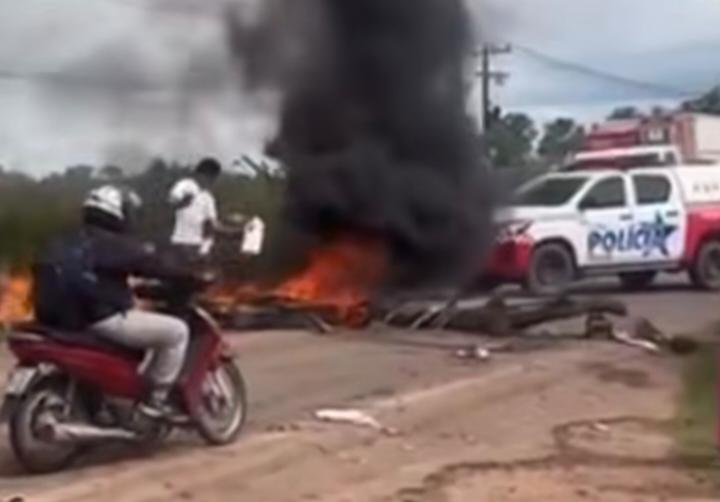 Homem em moto observando fogueira com fumaça preta bloqueando estrada e viatura policial ao fundo.