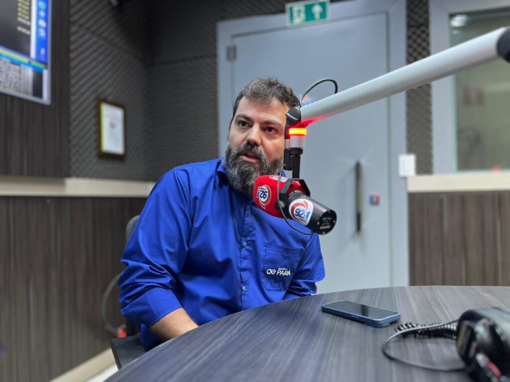 Homem barbudo de camisa azul em estúdio de rádio, próximo a um microfone profissional.