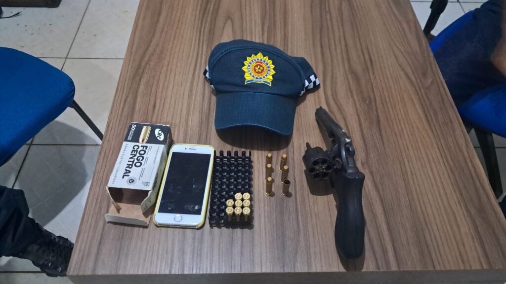 Boné com brasão, revólver, munições e celular sobre mesa de madeira.