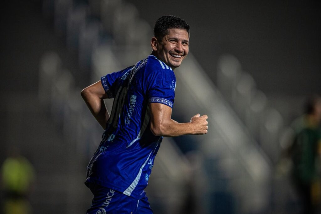 Jogador de futebol sorrindo em campo com camisa azul.