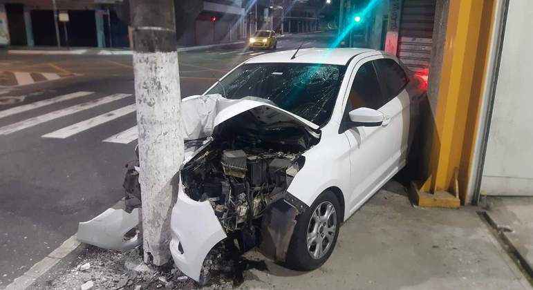 Carro branco acidentado contra poste e parede de loja.