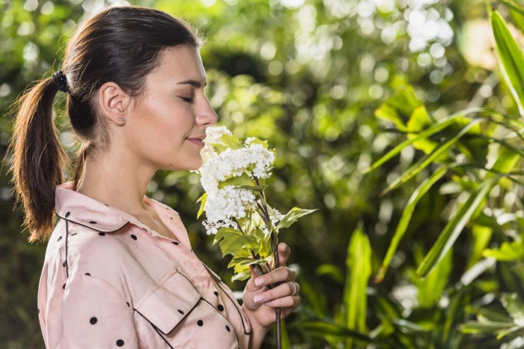 Mulher com olhos fechados cheirando flores brancas em um ambiente natural.