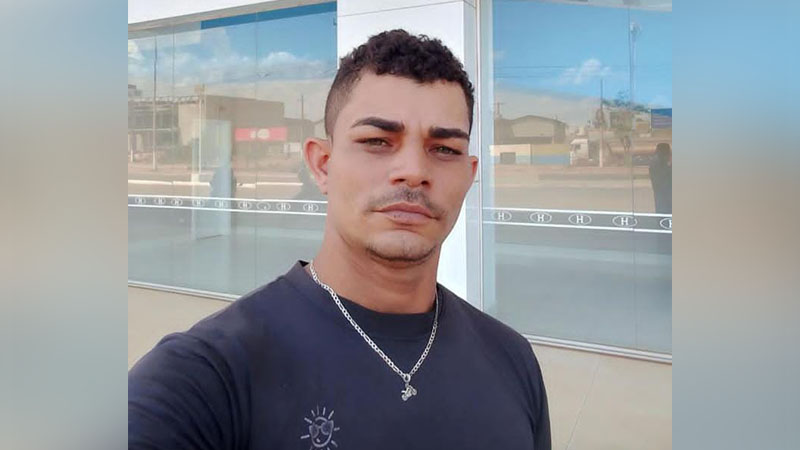 Homem com cabelo cacheado e bigode, vestindo camiseta preta e colar, em frente a um prédio de vidro.