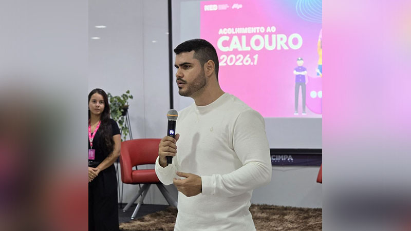 Homem falando ao microfone em evento de acolhimento a calouros, com 'ACOLHIMENTO AO CALOURO 2026.1' projetado ao fundo.
