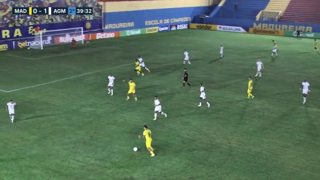 Partida de futebol com jogadores em campo, um deles driblando a bola.