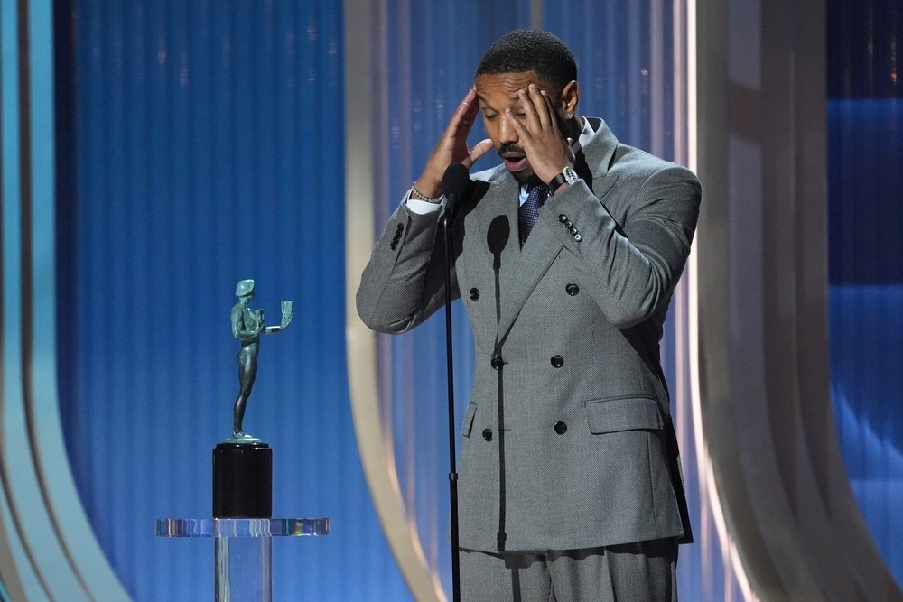 Michael B. Jordan emocionado no palco de uma cerimônia de premiação, mãos na cabeça, ao lado de um troféu SAG.