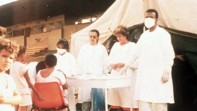 Grupo de profissionais de saúde com máscaras e crianças em um acampamento ou clínica provisória.