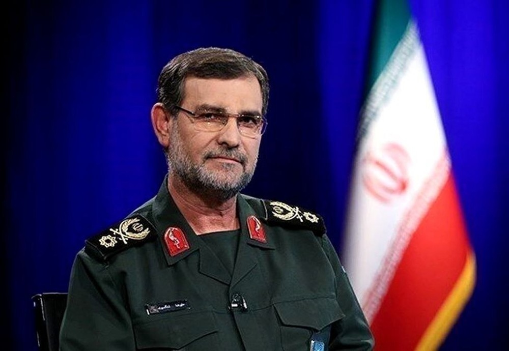 Homem em uniforme militar com bandeira iraniana ao fundo.