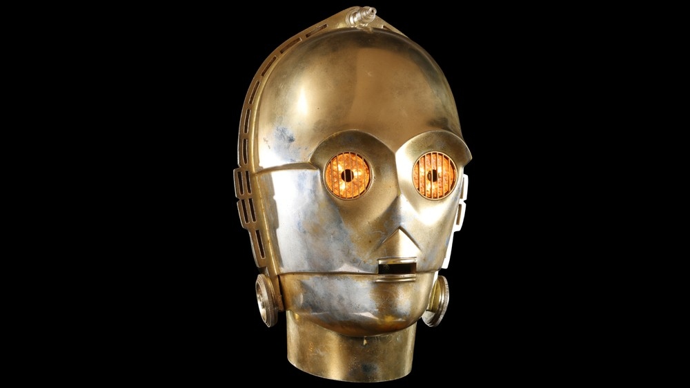 Cabeça do C-3PO, robô dourado de Star Wars, com olhos brilhantes em fundo preto.