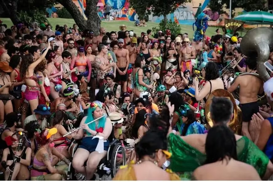 Multidão animada em um bloco de carnaval ao ar livre, com uma musicista em cadeira de rodas tocando flauta.