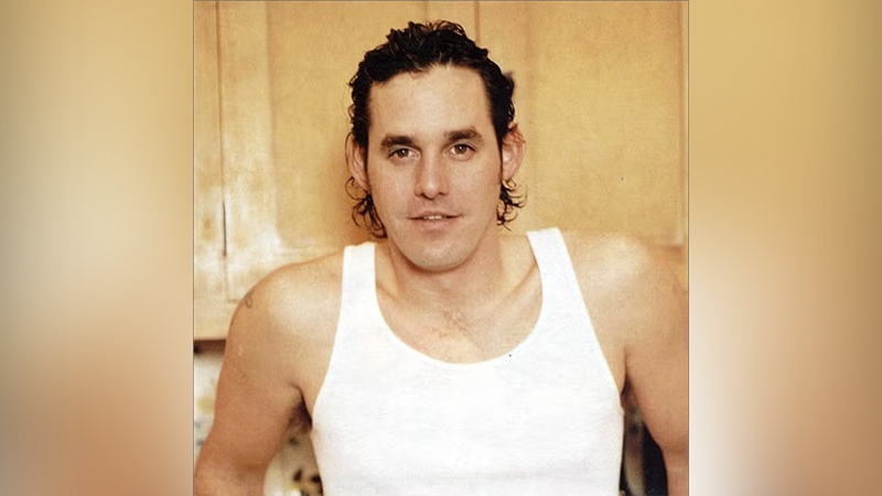 Homem de cabelo escuro encaracolado e regata branca olhando para a câmera.