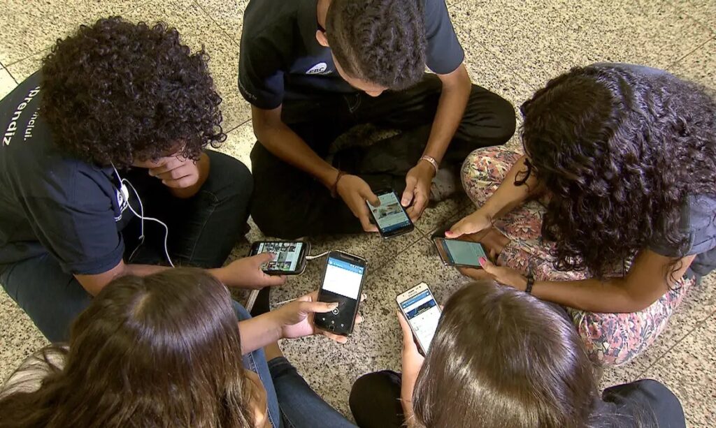 Grupo de jovens sentados no chão, cada um olhando atentamente para seu smartphone.