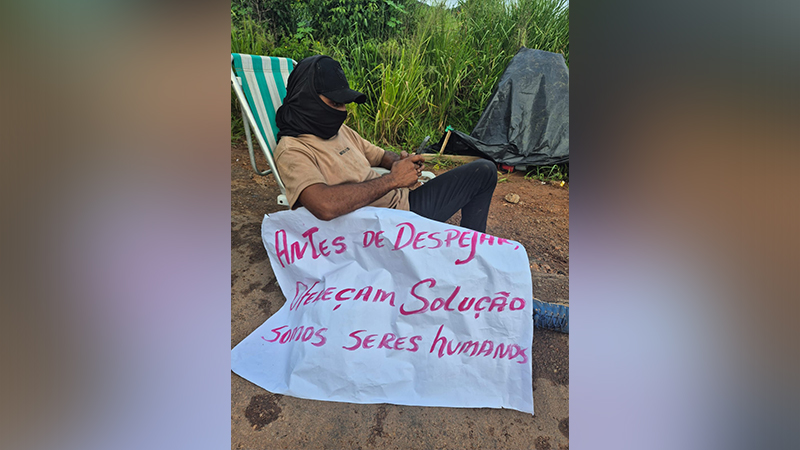 Homem mascarado sentado em cadeira de praia com cartaz "ANTES DE DESPEJAR, PEÇAM SOLUÇÃO SOMOS SERES HUMANOS".