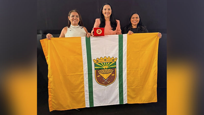 Três mulheres sorridentes segurando uma bandeira municipal e um prêmio em uma caixa vermelha.