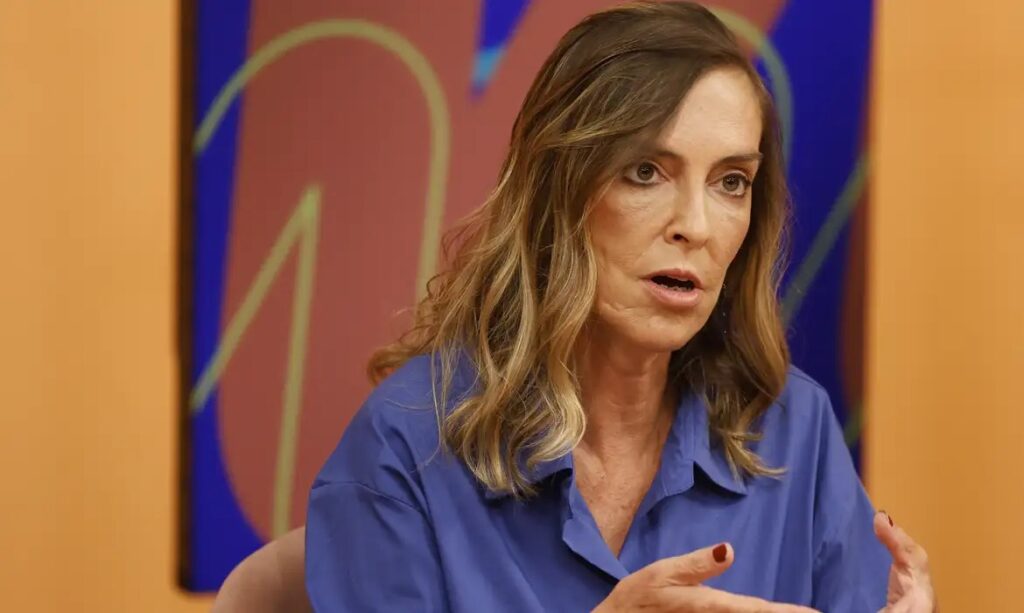 Mulher de cabelos castanhos claros e blusa azul, falando em um evento.