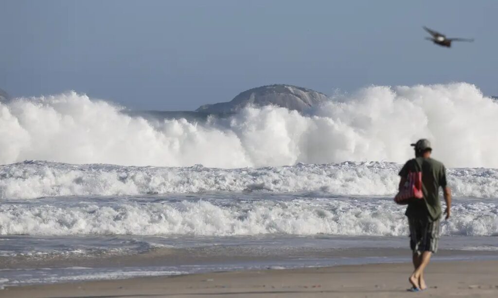 Ondas grandes e brancas quebrando na praia com uma ilha rochosa ao fundo sob céu azul.