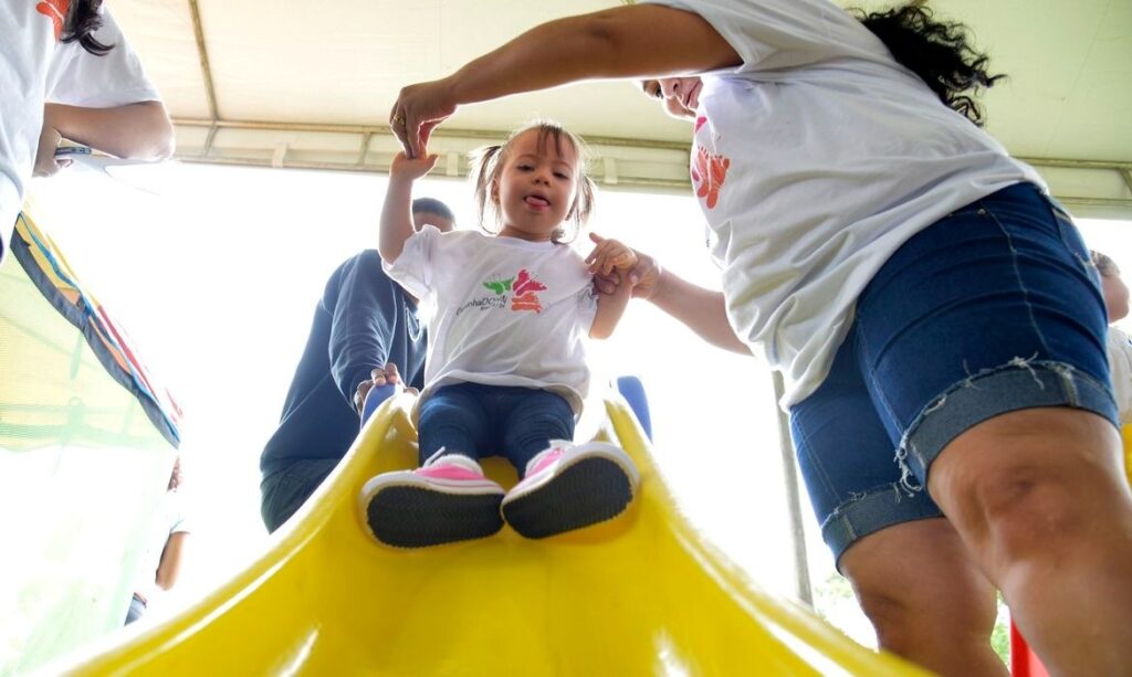 Menina com síndrome de Down em um escorregador amarelo, sendo ajudada por um adulto.
