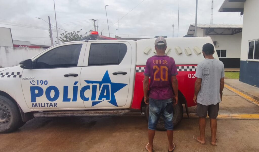 Viatura policial branca com detalhes vermelhos e azuis, e dois homens de costas à frente.