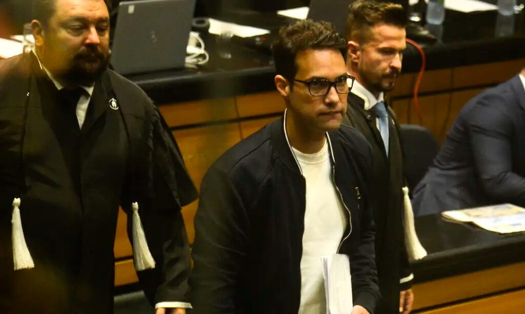 Homem de óculos e jaqueta bomber escura em ambiente formal, possivelmente um tribunal.