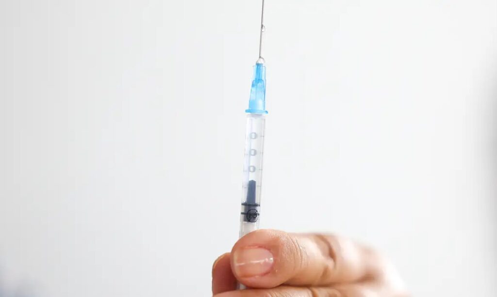 Mão segurando uma seringa com agulha e uma gota de líquido na ponta.