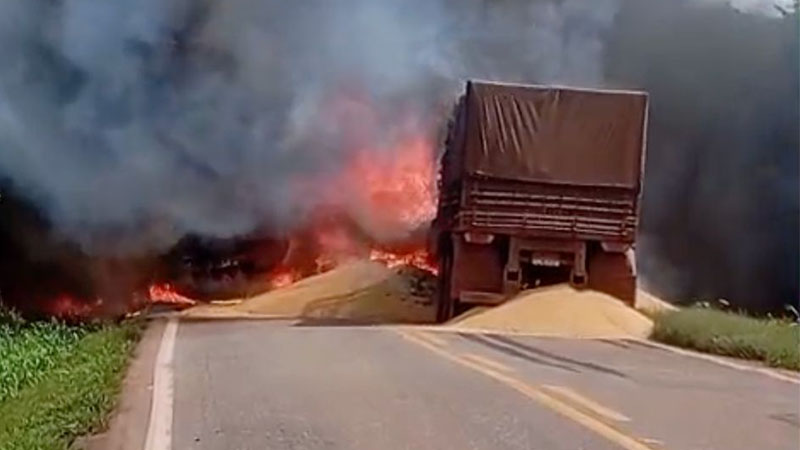 Caminhão graneleiro em chamas com carga de grãos derramada na estrada.