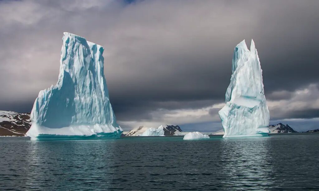 Dois enormes icebergs flutuando em águas escuras sob um céu nublado, com montanhas de gelo ao fundo.