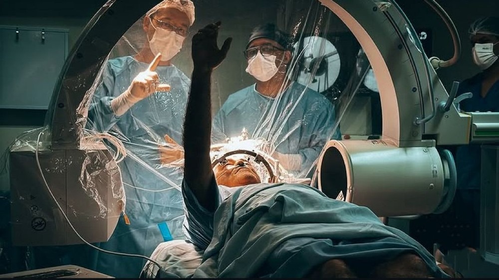 Paciente com capacete estereotáxico e braço levantado durante cirurgia, com dois médicos ao fundo.