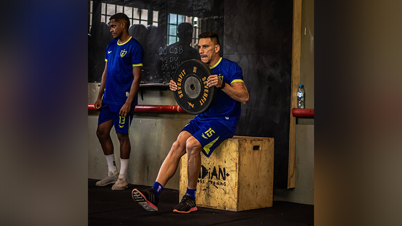 Homem em camisa azul e shorts, segurando um peso, sentado em um caixote de madeira em um ginásio.