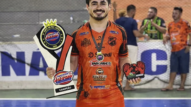 Homem sorridente em uniforme de futsal laranja segurando troféu de campeão e prêmio de melhor goleiro.