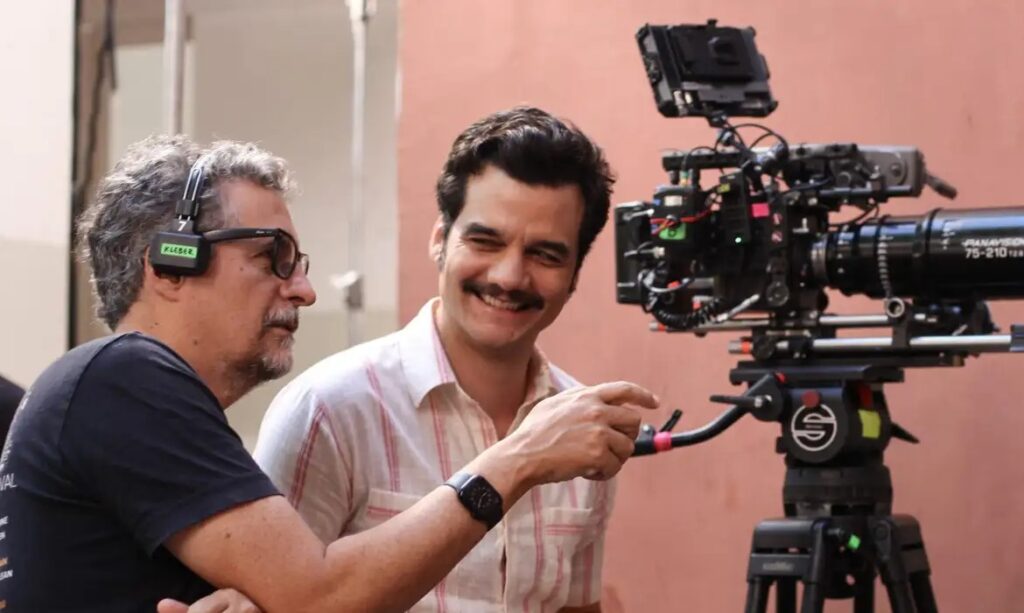 Wagner Moura sorrindo e apontando em um set de filmagem, ao lado de um colega e uma câmera profissional.