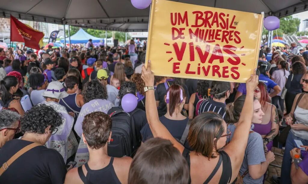 Uma multidão de mulheres em um protesto, com um cartaz amarelo levantado escrito 'Um Brasil de Mulheres Vivas e Livres'.