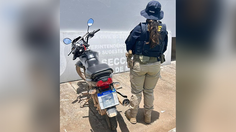 Policial da PRF de costas ao lado de uma motocicleta preta com placa brasileira, em frente a um prédio cinza.