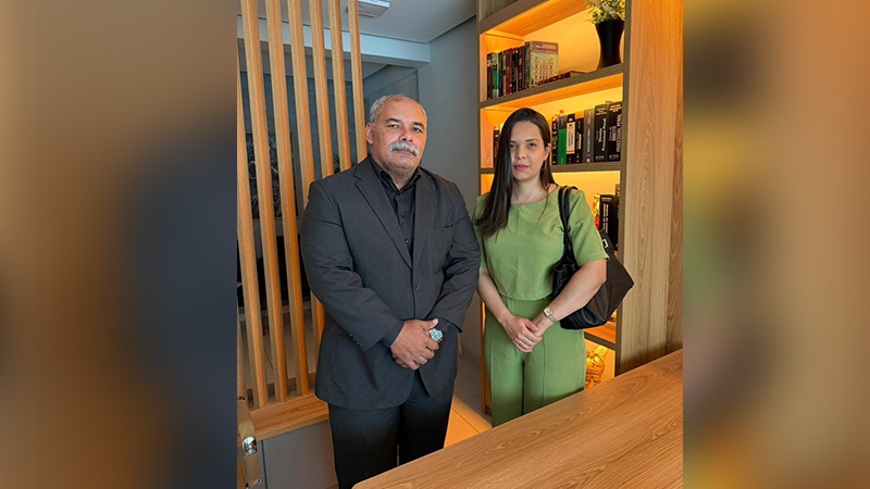 Um homem de terno preto e uma mulher de conjunto verde posam em um escritório com estante de livros.