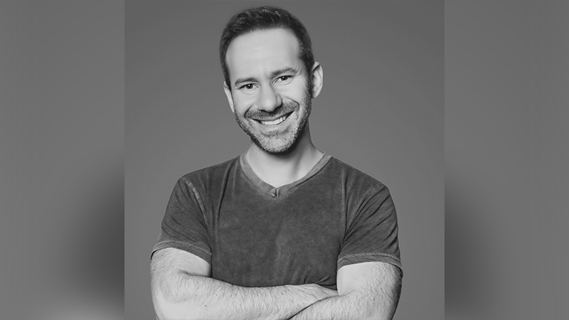 Homem sorridente com barba e braços cruzados em foto preto e branco.
